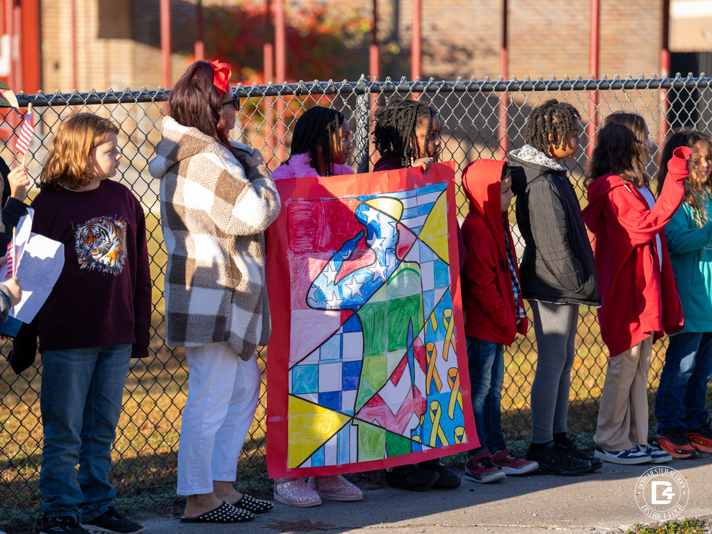 A line of students stands against a fence holding a brightly colored, handmade patriotic poster during the parade.
