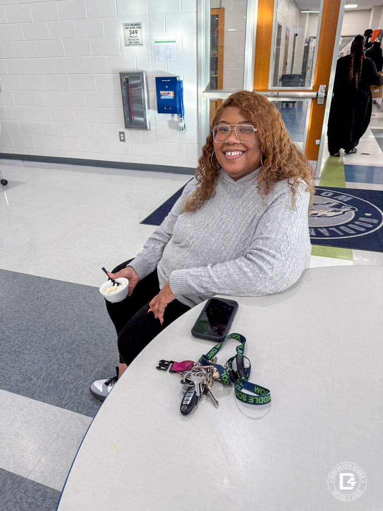 A woman in a gray sweater sits at a round table holding a cup of ice cream and smiling. Her keys and phone rest on the table in front of her in a bright school hallway.