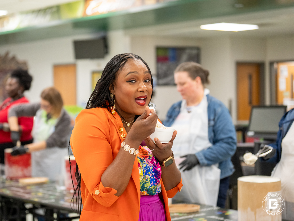 A woman in an orange blazer and colorful top smiles playfully while eating ice cream. Staff members serve ice cream in the background.