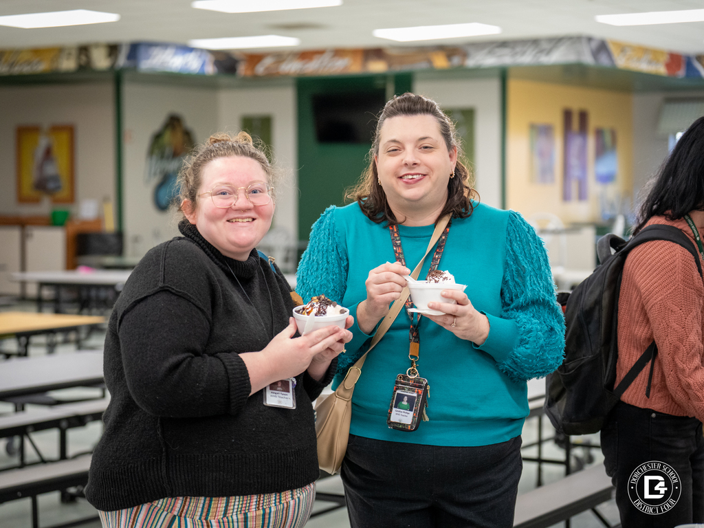 Two women stand in a cafeteria holding bowls of ice cream and smiling at the camera.