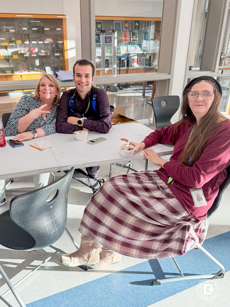 Three staff members sit together at a long table, smiling with bowls of ice cream. Sunlight comes through large windows behind them.