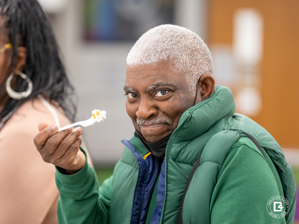A man in a green puffer jacket smiles at the camera while holding a spoonful of ice cream.