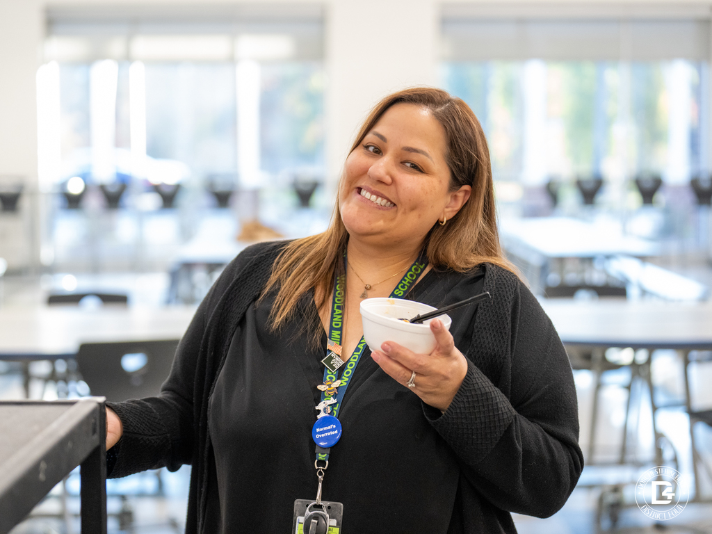 A woman wearing a black sweater and a Woodland Middle School lanyard smiles while holding a bowl of ice cream in the cafeteria.