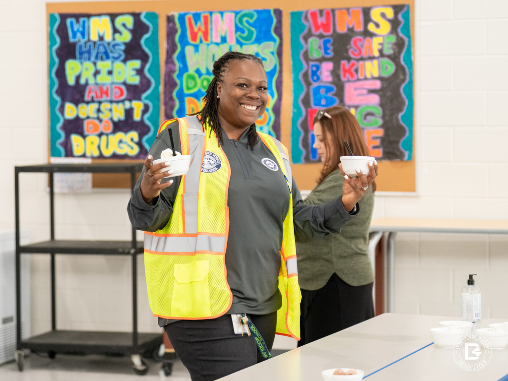 A staff member in a yellow safety vest holds two bowls of ice cream and smiles while standing in front of colorful posters on the wall.