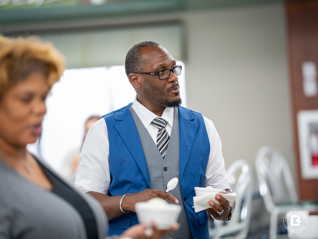 A man wearing a blue vest and tie holds a bowl of ice cream while talking with others in the cafeteria.