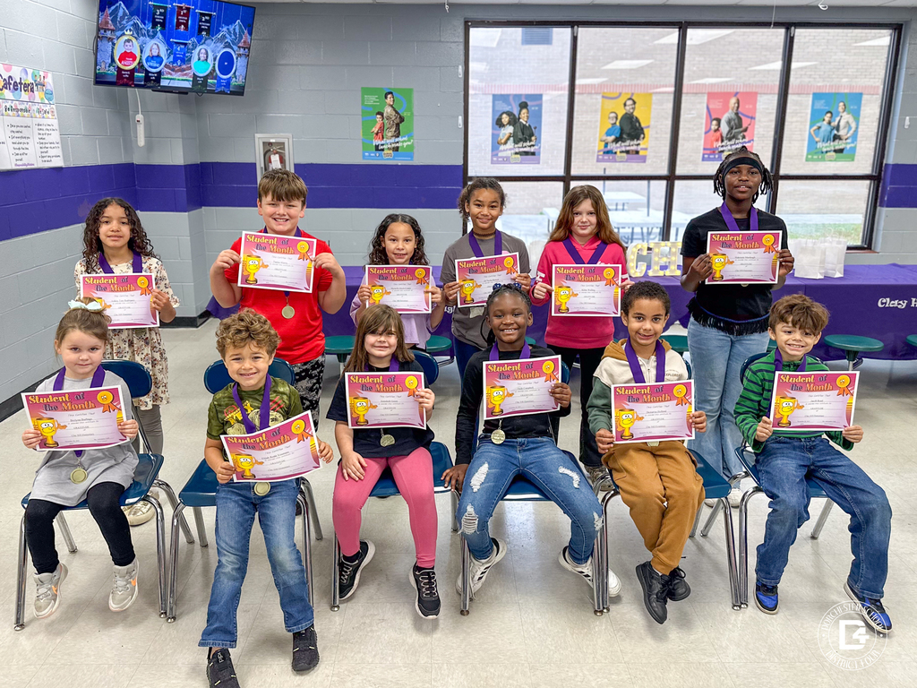 A group of Clay Hill Elementary students sit in two rows holding “Student of the Month” certificates and wearing medals. Posters and windows line the wall behind them.