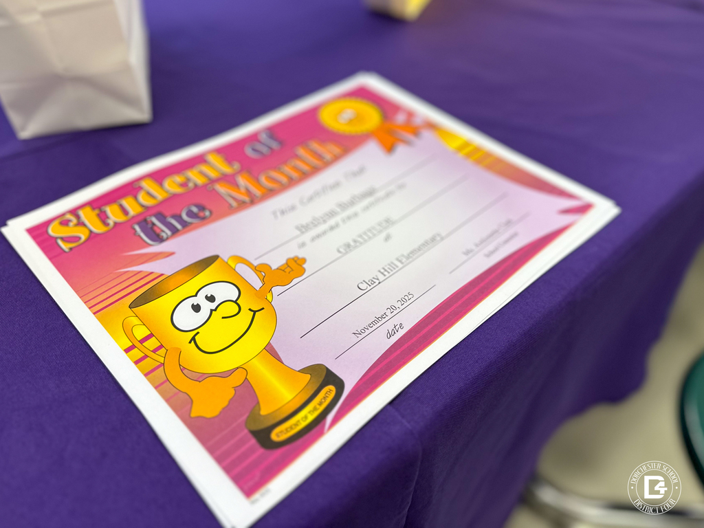 A close-up of a colorful “Student of the Month” certificate with a smiling trophy graphic, placed on a purple tablecloth.