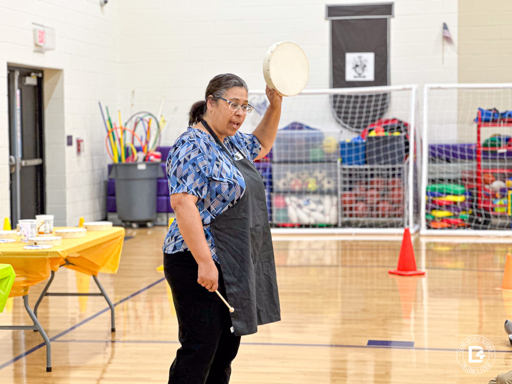 Ms. Tammy Leach holds up a hand drum and speaks to students in the gym, with painting supplies set out on tables nearby.