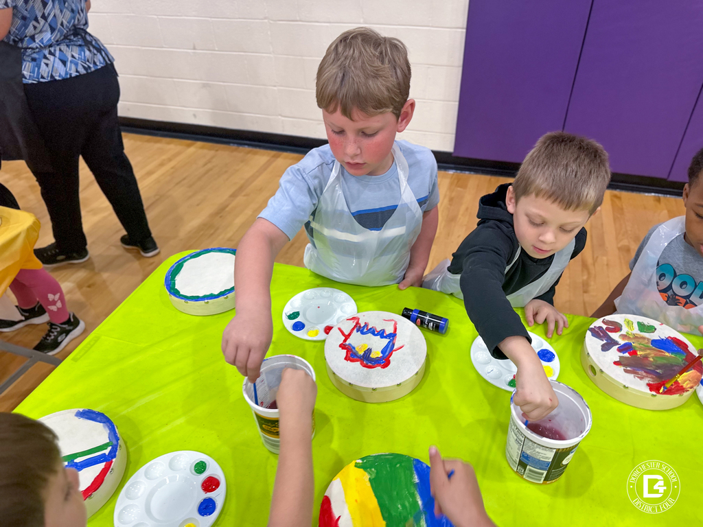 Two boys paint their hand drums at a bright green table, dipping their brushes into paint cups as they work on colorful designs.