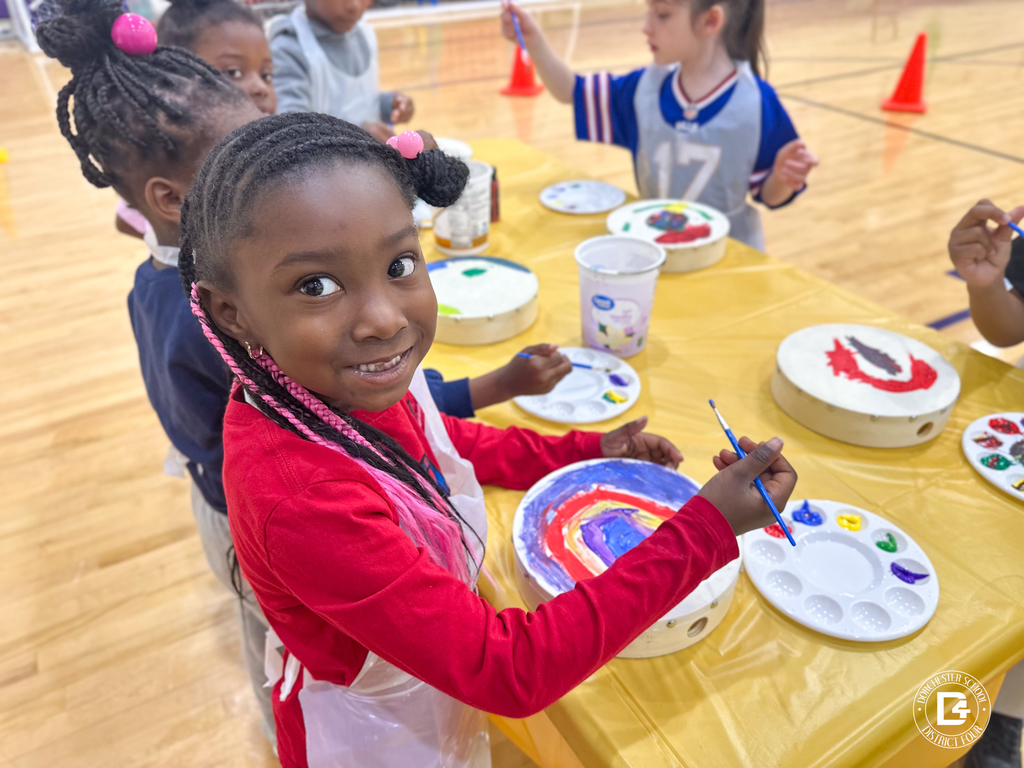 A young girl smiles at the camera while painting a hand drum at a yellow-covered table with other students.