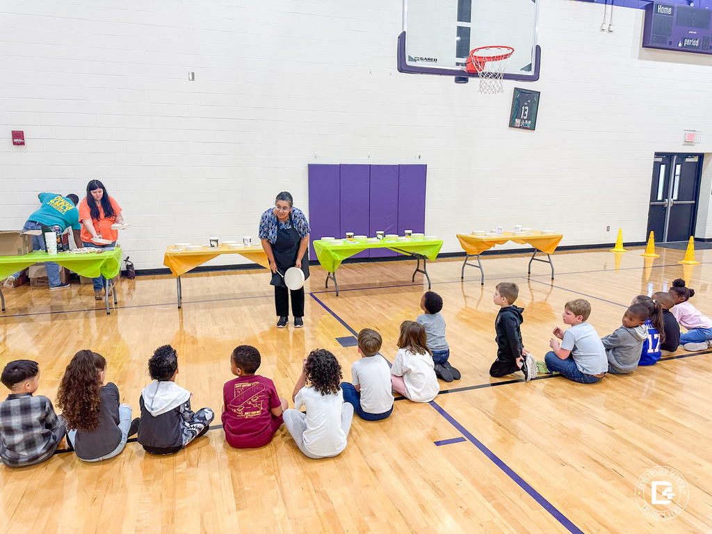 Students sit on the gym floor facing Ms. Tammy Leach, who stands holding a hand drum while two volunteers prepare materials at tables behind her.