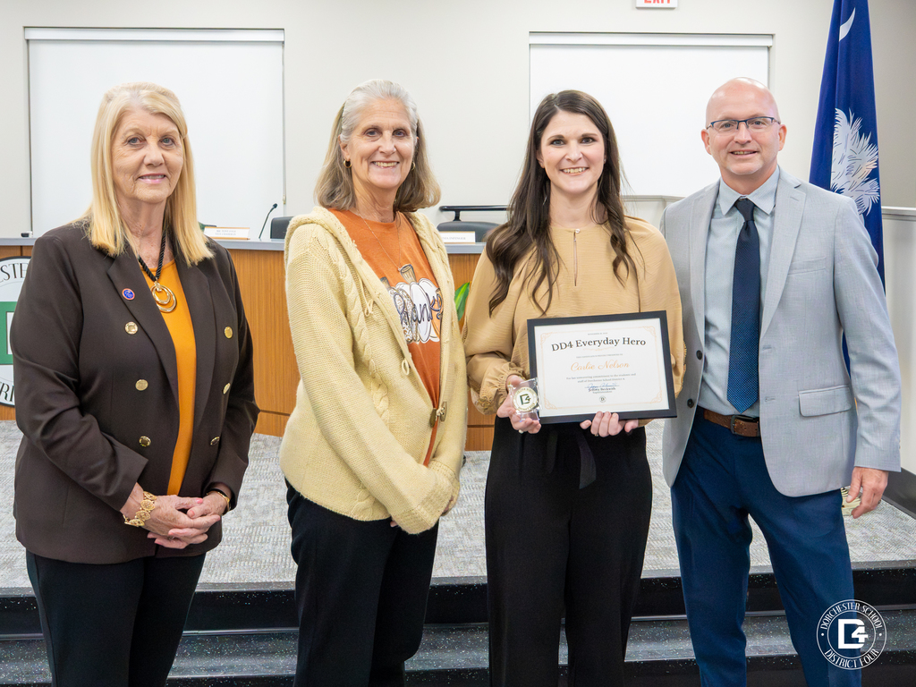 Carlie Nelson stands with three district leaders in the DD4 board room, holding her DD4 Everyday Hero certificate and coin while smiling toward the camera.