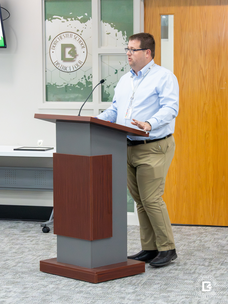 A man stands at a podium in the DD4 board room giving a presentation. He wears glasses, a light blue dress shirt, khaki pants, and a district badge as he speaks into the microphone.