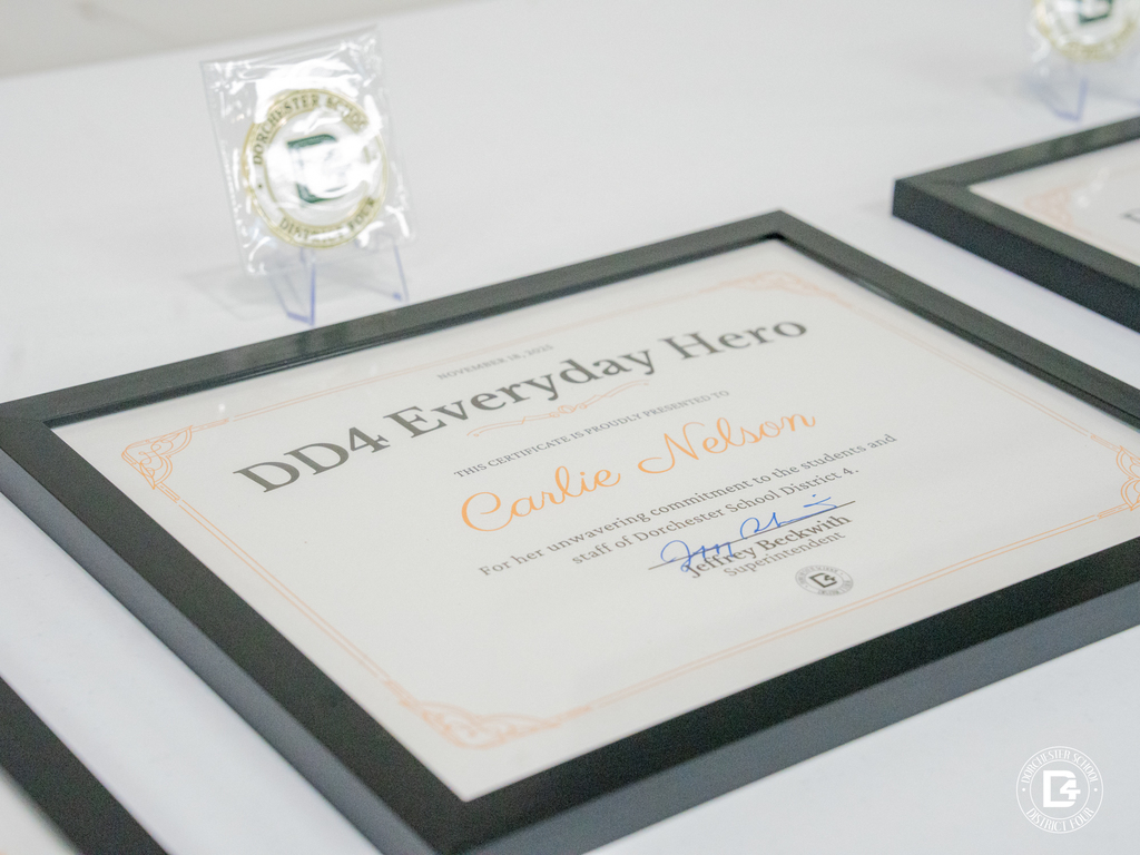 A framed DD4 Everyday Hero certificate for Carlie Nelson lies on a white table with a DD4 challenge coin displayed upright behind it.