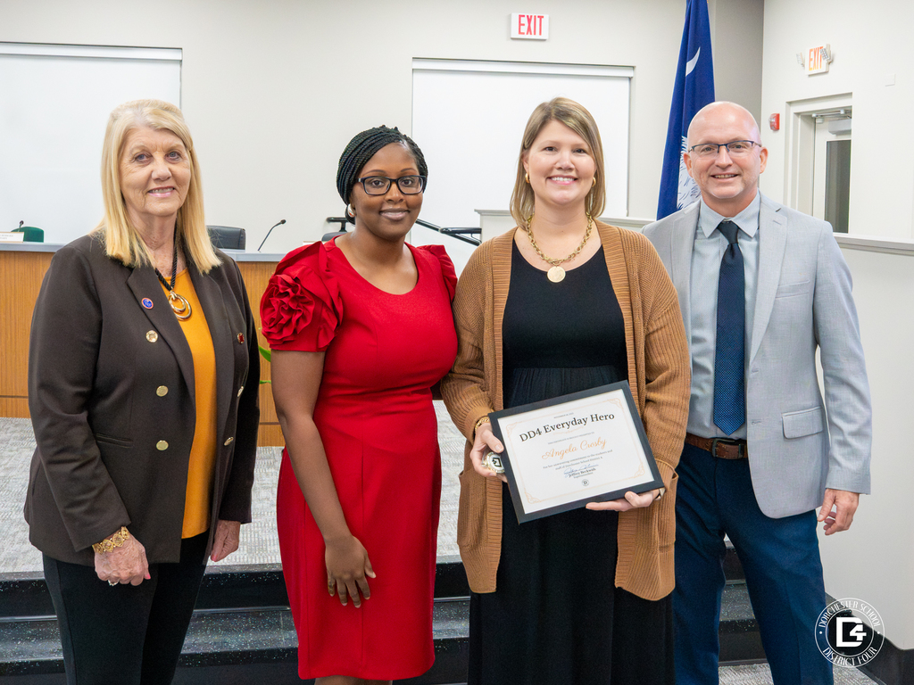 Four adults stand together at the DD4 board room. Angela Crosby holds her DD4 Everyday Hero certificate and coin while smiling, with two district leaders and a board member standing beside her.