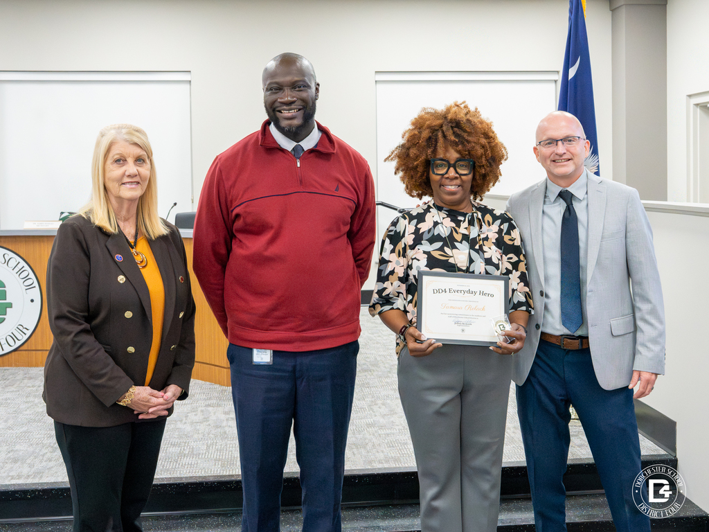 Tamara Rolack stands with three district leaders in the DD4 board room. She holds her DD4 Everyday Hero certificate and challenge coin while smiling toward the camera.