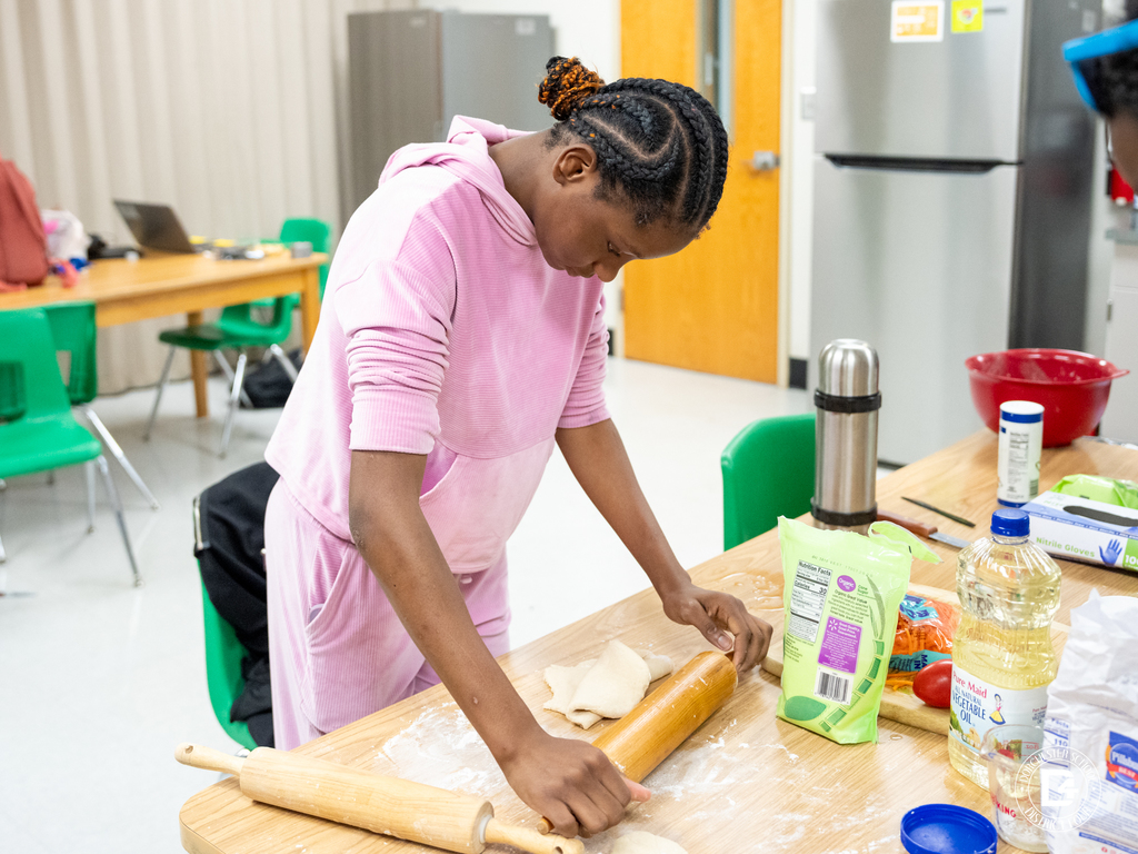 A student in a pink tracksuit uses a rolling pin to flatten dough on a flour-covered table, surrounded by cooking supplies and utensils.