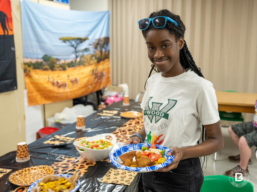 A student wearing sunglasses on her head holds a blue paper plate filled with Kenyan breakfast items while standing beside a table decorated with safari-themed plates and cups.