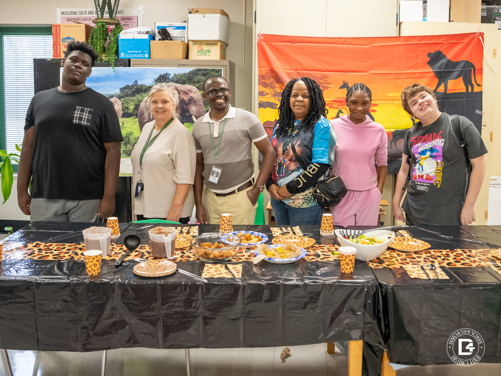 A group photo of six Woodland High School students and staff standing behind a long table covered with black and leopard print decor, with plates of Kenyan breakfast foods displayed. A colorful safari-themed backdrop with silhouettes of animals hangs behind them.