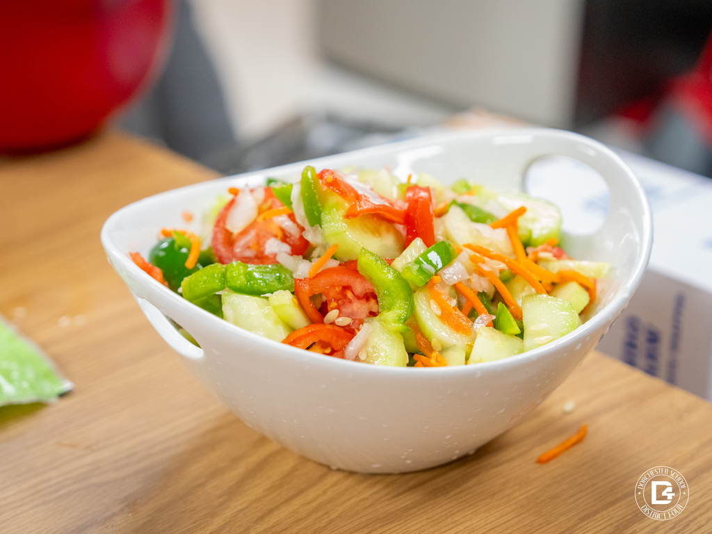 A white bowl filled with a bright, fresh salad of cucumbers, tomatoes, bell peppers, onions, and shredded carrots.