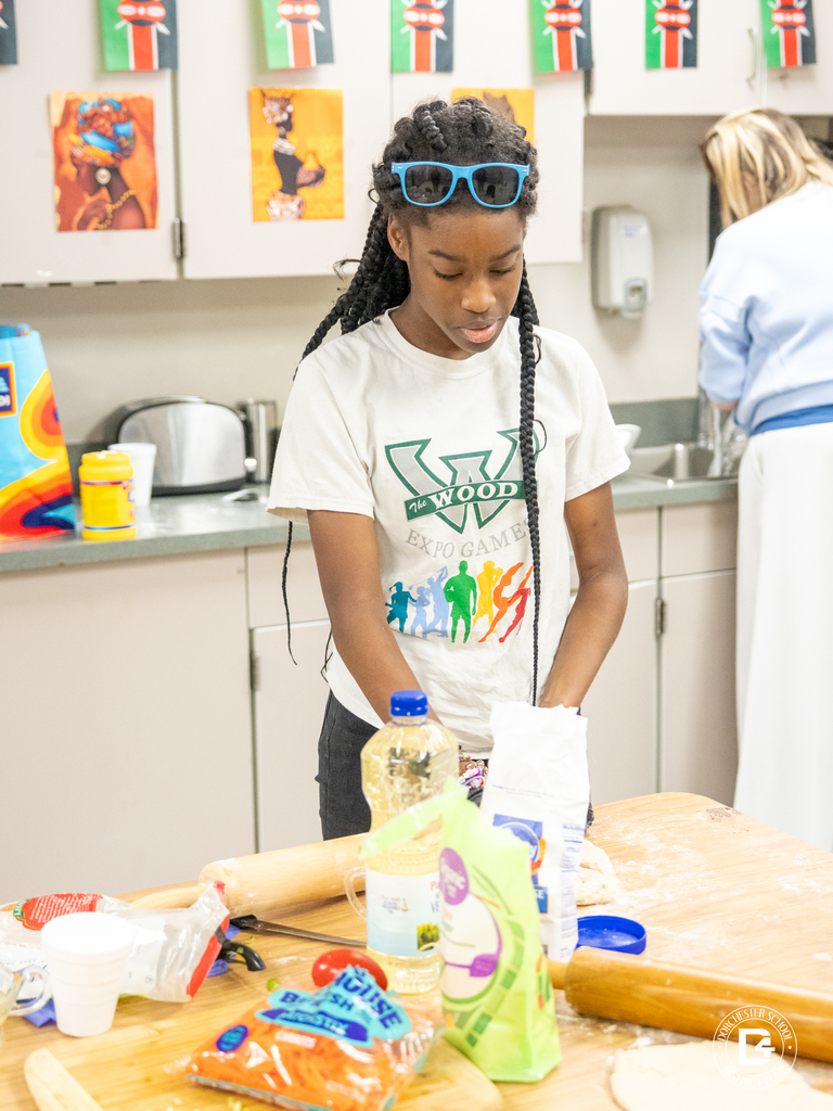 A student wearing a Woodland Expo Games shirt kneads dough at a table covered with baking ingredients and utensils.