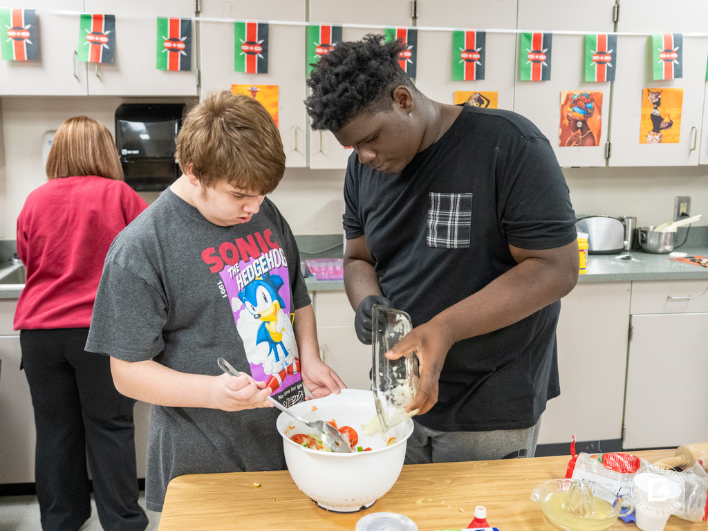 Two students work together at a table, one pouring chopped onions while the other mixes vegetables for a salad. More Kenyan flags hang behind them.