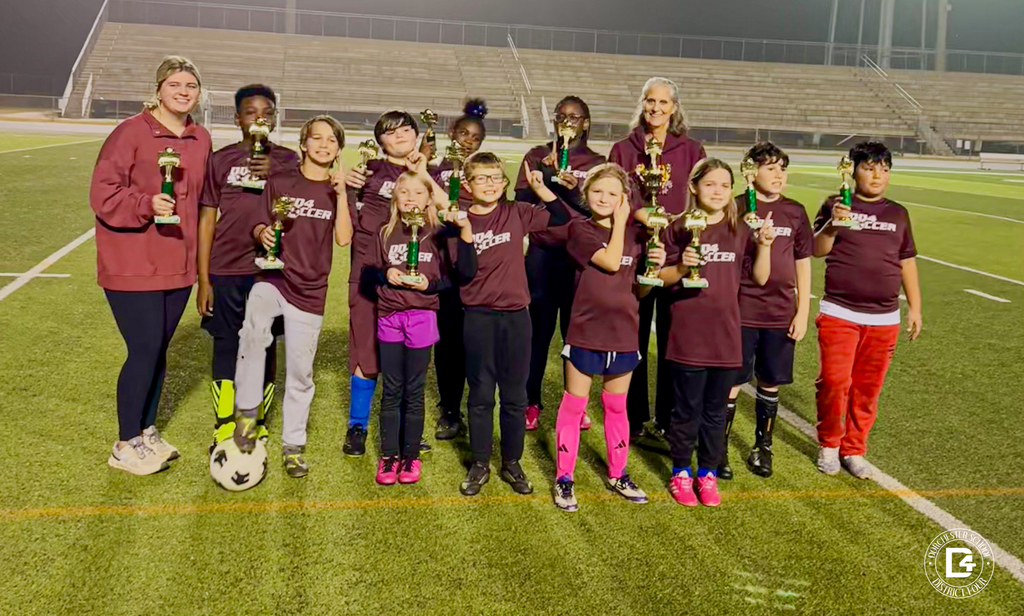 A youth soccer team stands on a football field at night, smiling and holding gold trophies. The group includes about a dozen children wearing maroon DD4 Soccer shirts along with two adult coaches, one on each side. The kids stand in a row on the turf with bleachers in the background, and a soccer ball rests near the feet of one player.