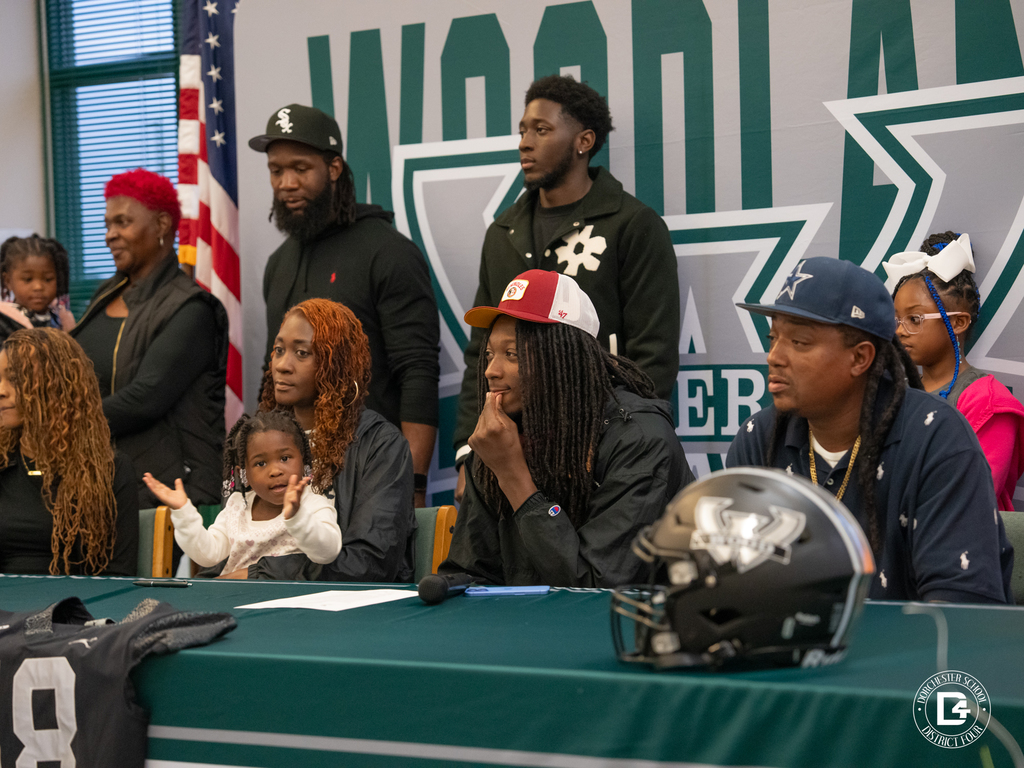 Jaemin Pinckney sits at a signing table surrounded by family, with a Woodland backdrop and his number 18 jersey displayed in front.