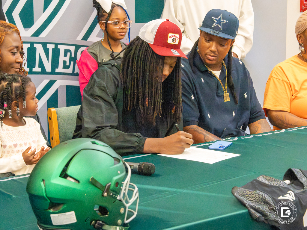 Jaemin Pinckney signs his letter of intent at the table while family looks on; a green Woodland helmet sits nearby.