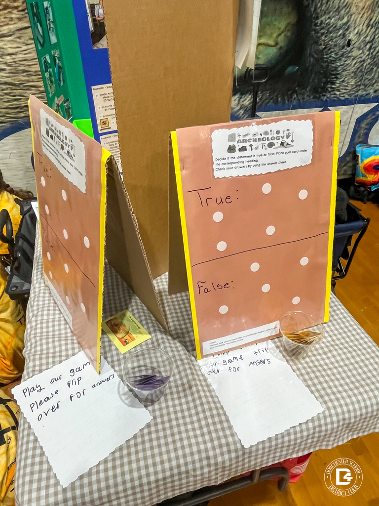 Students and coaches in yellow Tiger Bots shirts wait around a robotics table inside a busy gymnasium as multiple teams compete. Spectators sit in the bleachers behind them.