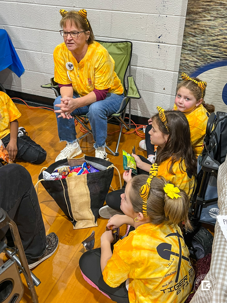 Tiger Bots students sit on the gym floor eating snacks while a coach reads from a booklet. Other teams and tables fill the background.
