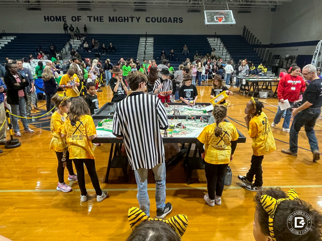 Three Tiger Bots students stand with a referee reviewing a score sheet at the competition table. The students wear yellow tie-dye shirts with Tiger Bots printed on the back.