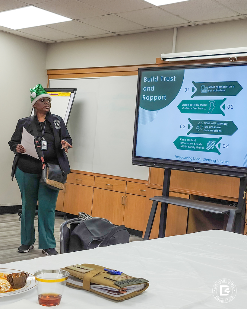 A presenter stands beside a screen reading “Build Trust and Rapport,” sharing mentoring best practices with community members seated at tables during a Dorchester School District Four mentoring program meeting.