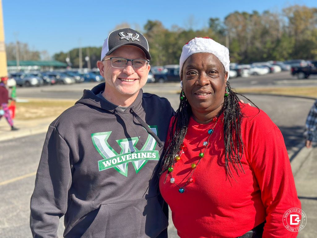 A staff member and community volunteer pose together outdoors during Christmas at DD4, with the school campus visible behind them.