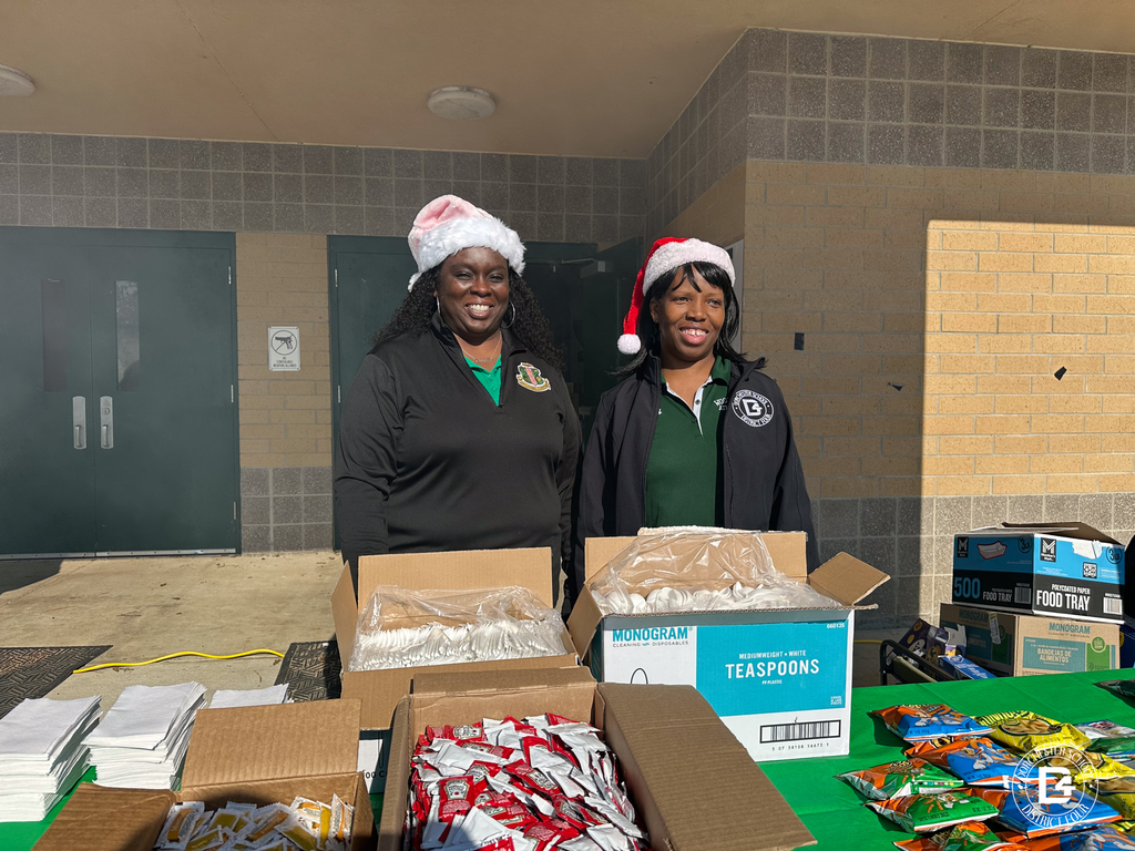 Two volunteers wearing Santa hats stand behind tables stocked with napkins, utensils, and snacks, helping distribute food to attendees.