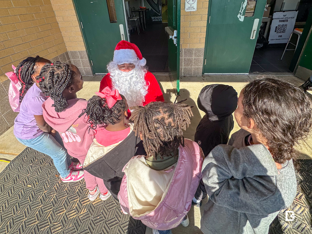 Santa Claus kneels and speaks with a group of young children gathered around him outside Woodland High School.