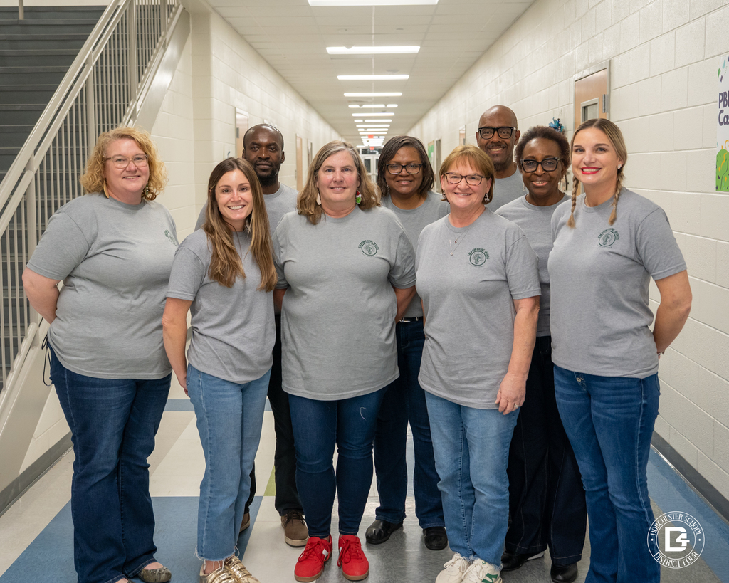 Group of Dorchester School District Four staff members standing shoulder to shoulder in a school hallway, smiling at the camera while wearing matching gray district T-shirts and jeans.