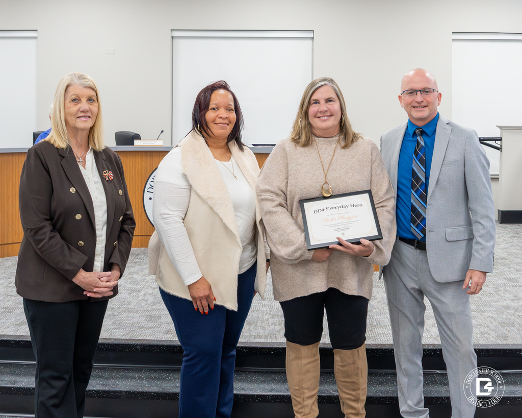 Paula Huggins, a CHES teacher, holds her DD4 Everyday Hero certificate while standing with district leadership in front of the board room, captured in a proud moment following her recognition.