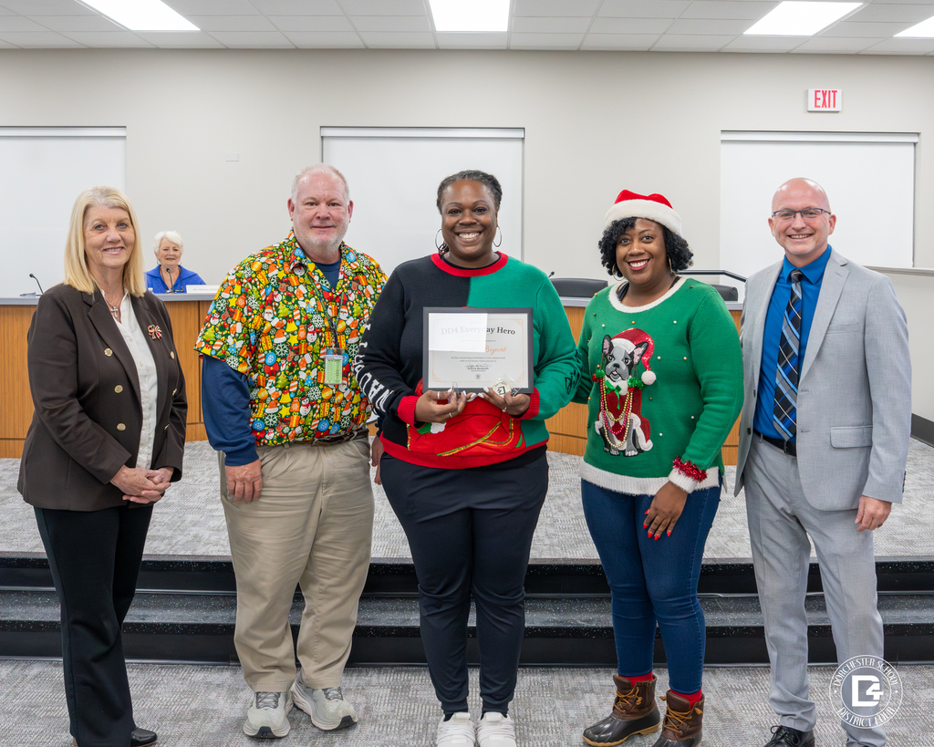 Latorsha Bryant, assistant principal at Woodland Middle School, holds her DD4 Everyday Hero certificate while standing with colleagues and district leaders, several dressed in festive holiday attire, marking her recognition during the December board meeting.