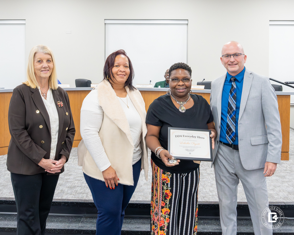 Lakisha Pigatt, a CHES interventionist, stands centered holding her DD4 Everyday Hero certificate and commemorative coin, surrounded by district leaders at the board meeting dais, all smiling during her recognition.