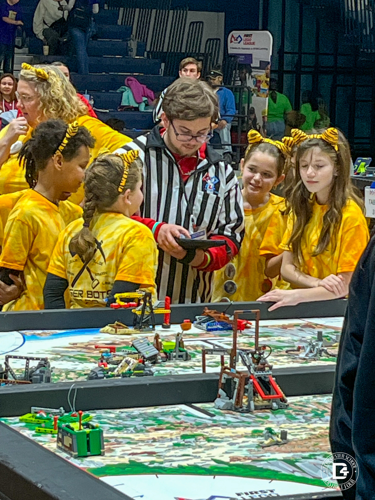A group of Williams Memorial Elementary Tiger Bots students in yellow tie-dye shirts and tiger-ear headbands stand at a robotics competition table speaking with a referee in a black-and-white striped shirt who is reviewing a score sheet. LEGO mission models cover the table.