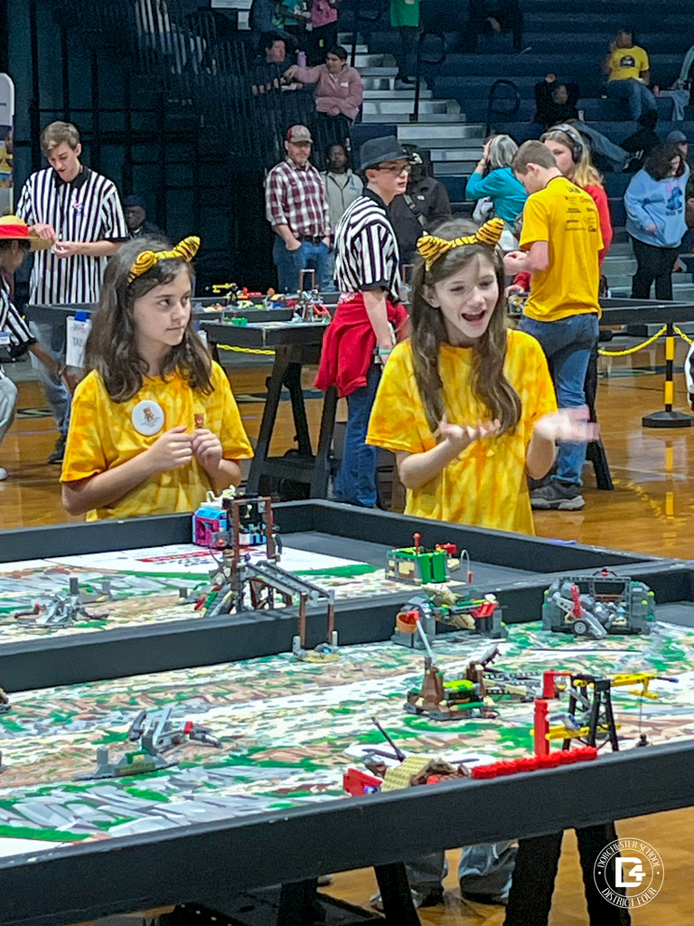 Two Tiger Bots students in yellow shirts and tiger-ear headbands stand at a FIRST LEGO League competition table, gesturing and watching their robot’s run while referees and other teams work in the background.