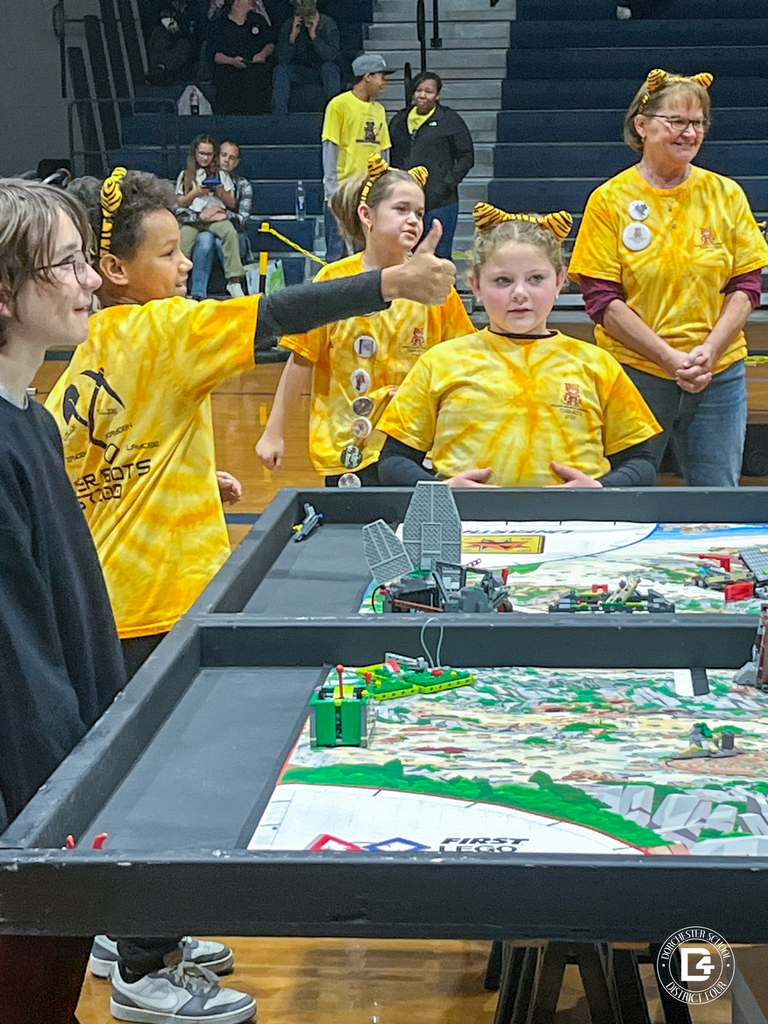 Tiger Bots team members gather around a competition table. One student gives a thumbs-up while another focuses on the robot game. Their coach stands nearby, smiling.