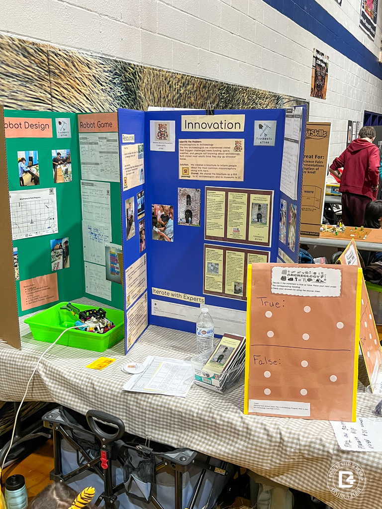 A tabletop display shows a handmade tri-fold board with a True or False archaeology game. Two cups of answer cards sit in front of the board on a checked tablecloth.
