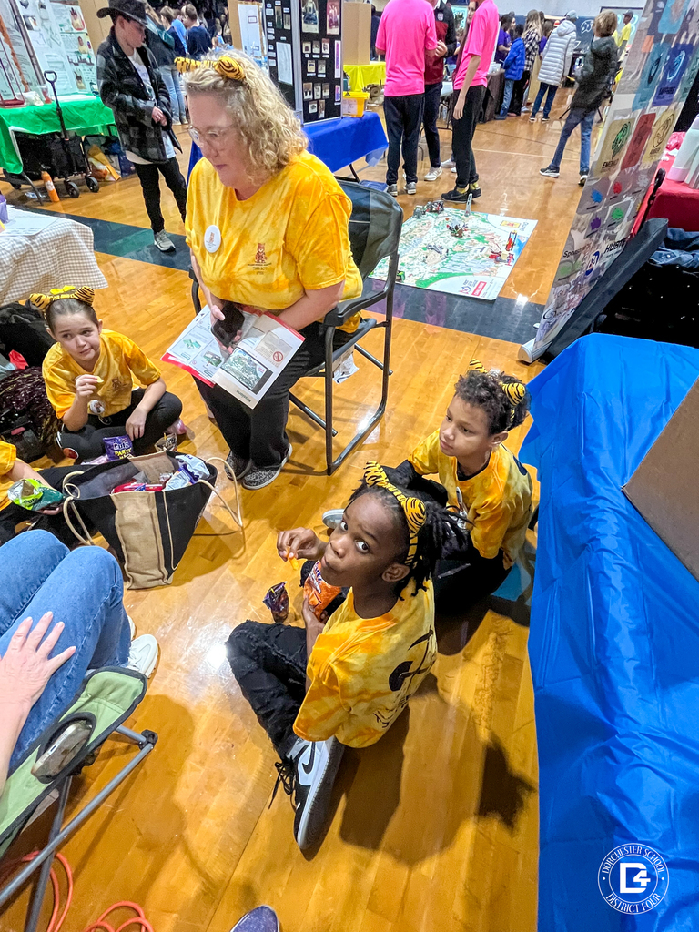 A full Tiger Bots research and innovation station is set up with tri-fold boards detailing robot design, the robot game, and an archaeology project. Handouts, LEGO pieces, and a water bottle sit on the table.