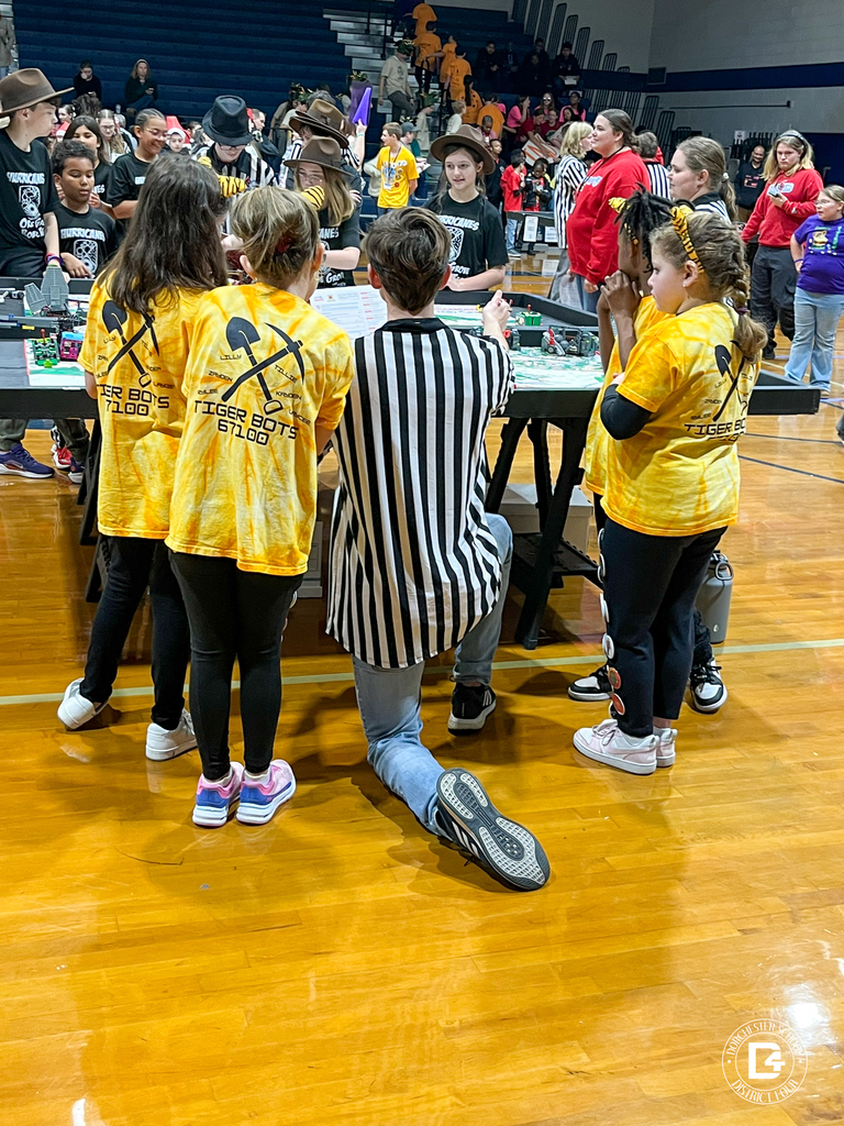 A Tiger Bots coach sits in a folding chair speaking with team members who are gathered around a tote full of snacks. Students wear yellow tie-dye shirts and tiger-ear headbands.
