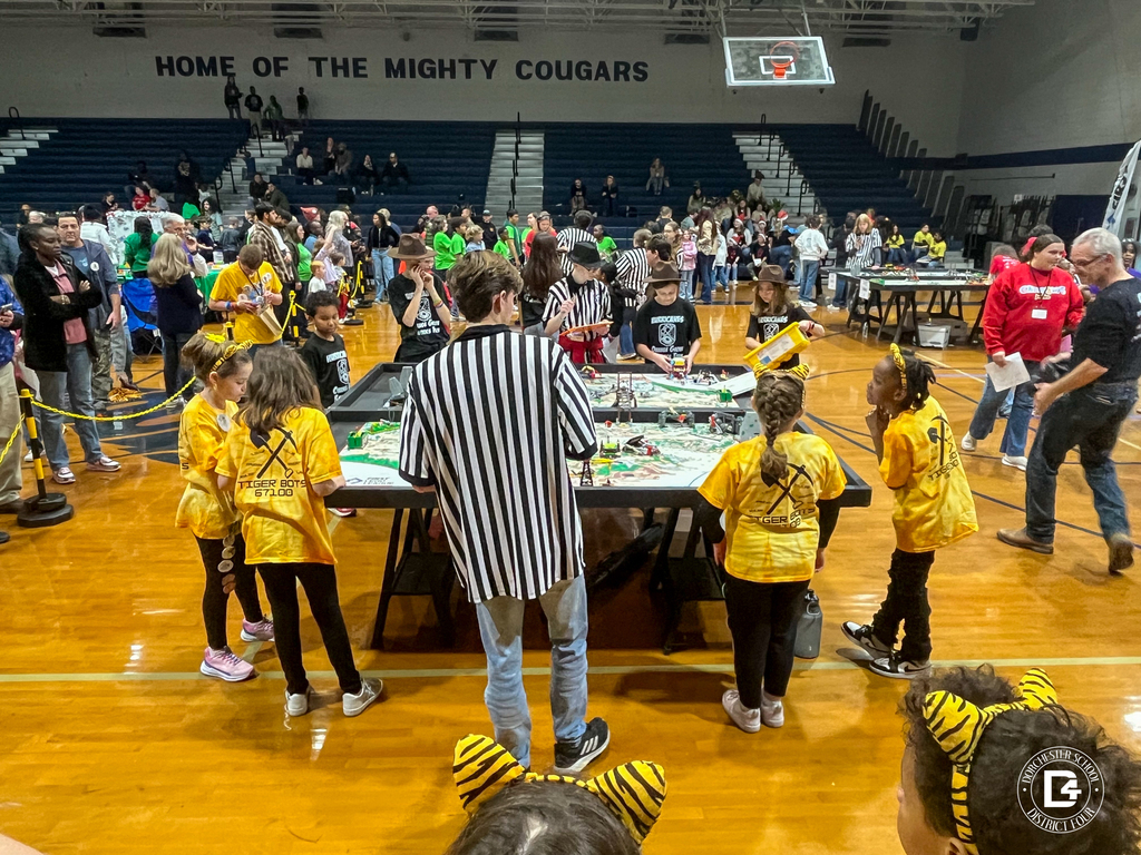 Three Tiger Bots students stand with a referee reviewing a score sheet at the competition table. The students wear yellow tie-dye shirts with Tiger Bots printed on the back.