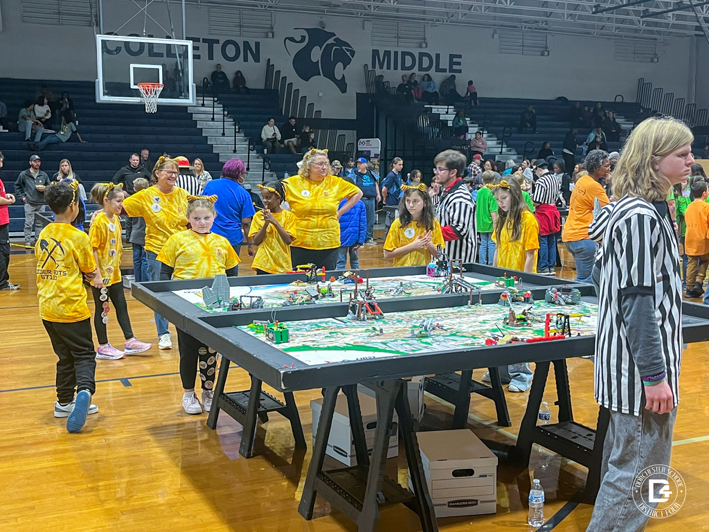 Tiger Bots students prepare for a match at a robotics table inside a crowded gym. Referees oversee multiple games while families and teams watch from the sidelines.