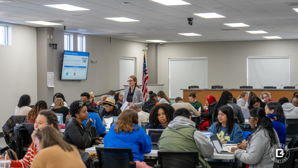Educators collaborate at tables while a presenter stands near a screen at the front of the room during a professional development workshop.