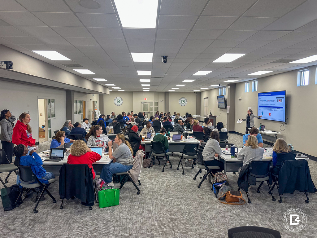 Wide view of educators seated at round tables during a professional development session, working on laptops and participating in group discussions in a large meeting room.